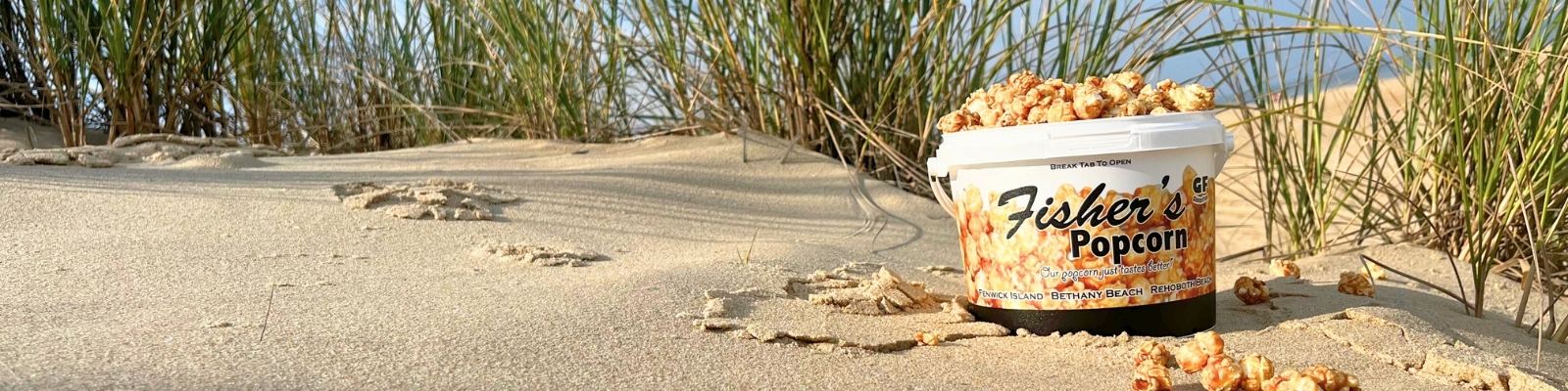 A tub of Fisher’s Popcorn sits on sandy dunes with scattered popcorn kernels, surrounded by beach grass under a clear sky.