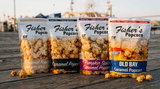 Four bags of Fisher’s Popcorn in different flavors, including caramel, pumpkin spice, and Old Bay, displayed on a wooden boardwalk with a Ferris wheel in the background.