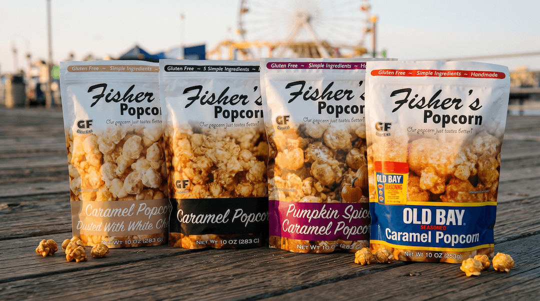Four bags of Fisher’s Popcorn in different flavors, including caramel, pumpkin spice, and Old Bay, displayed on a wooden boardwalk with a Ferris wheel in the background.