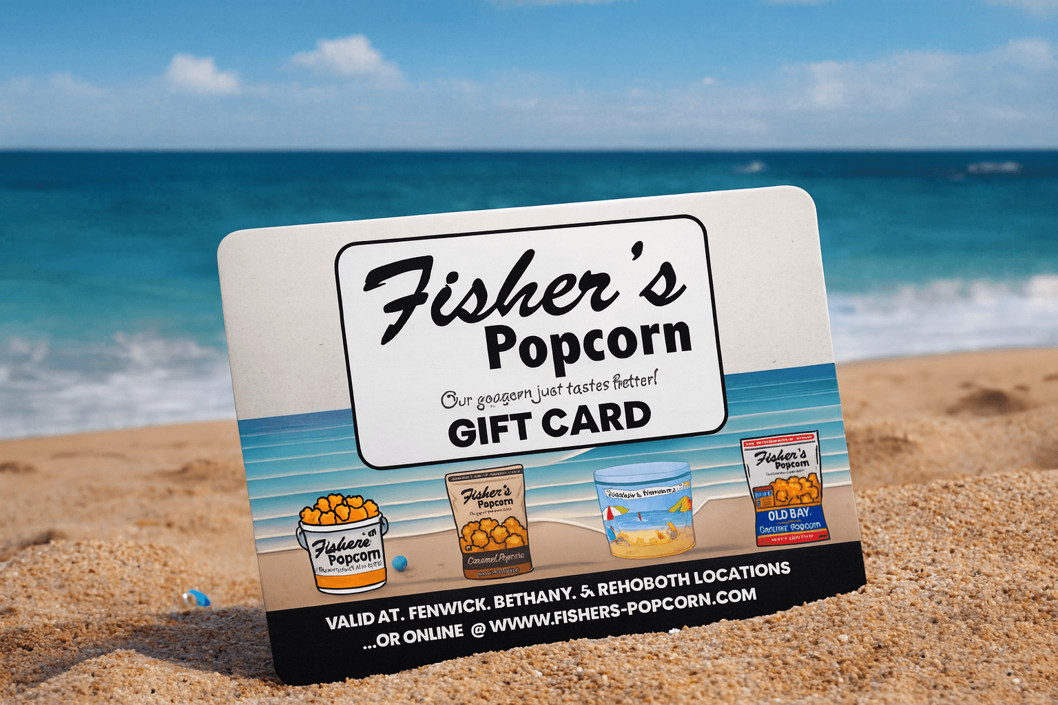 A Fisher’s Popcorn gift card is propped up on sandy beach with the ocean and blue sky in the background. The card shows popcorn tubs and promo text.