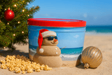 A festive popcorn tin on a sandy beach, decorated with a snowman in sunglasses and a Santa hat, sits beside popcorn, a Christmas tree, and gold ornaments with the ocean in the background.