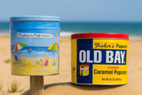 Two tins of Fisher’s Popcorn, one with a beach scene and one labeled “Old Bay Seasoned Caramel Popcorn,” sit on sandy beach with the ocean and blue sky in the background.