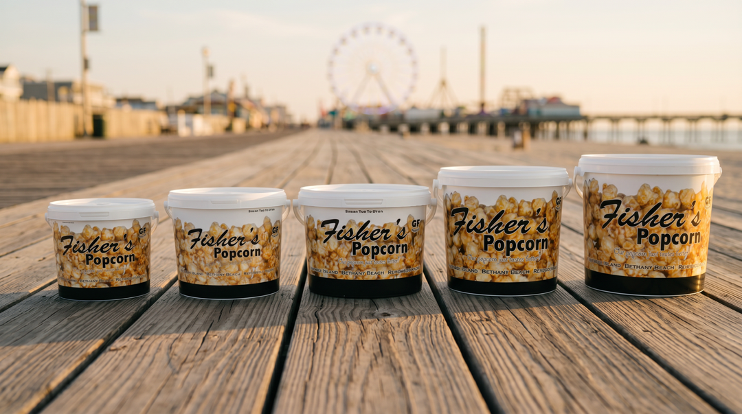Five containers of Fisher’s Popcorn in various sizes are lined up on a wooden boardwalk, with a ferris wheel and amusement park visible in the blurred background.