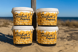 Four tubs of Fishers Popcorn are stacked on sandy ground at a beach, with clear blue sky and ocean in the background.