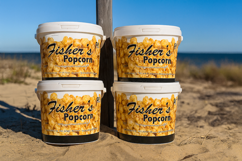 Four large tubs of Fisher’s Popcorn stacked on sandy ground near a beach, with blue sky and ocean visible in the background.