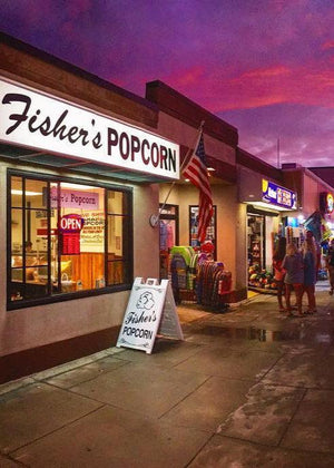 A street scene at dusk shows Fisher’s Popcorn shop with a lit sign and an American flag. People are gathered outside nearby stores under a purple and pink sky, and a bright OPEN sign is visible in the window.