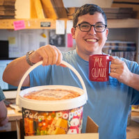 A smiling person wearing glasses and a blue shirt holds a large tub of Fisher’s Popcorn in one hand and a red mug labeled Mr. Claus in the other, standing in a cozy, cluttered room.