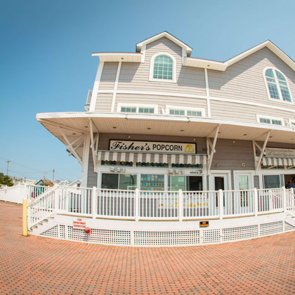 A light gray, two-story building with a sign reading Fishers Popcorn above a white-railed porch on a sunny day. The sidewalk and open sky are visible in front of the shop.