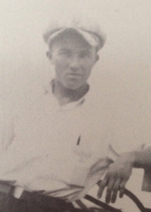 Black and white photo of a young man wearing a light-colored cap and shirt, looking at the camera with a neutral expression. He is seated and resting one arm on the back of a chair.