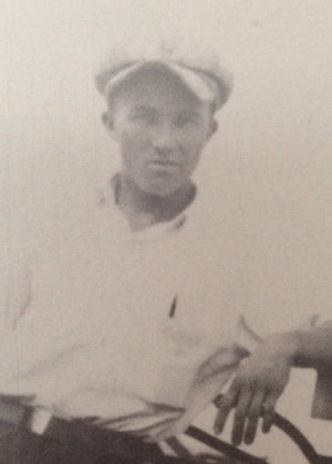 Black and white photo of a young man wearing a light-colored cap and shirt, looking at the camera with a neutral expression. He is seated and resting one arm on the back of a chair.