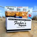 A BYOB 12 Pack of Large Bags sits on sand with a blue sky behind it, featuring a custom popcorn assortment in large resealable bags. The box notes the popcorn is gluten-free, handmade, and made with simple ingredients.