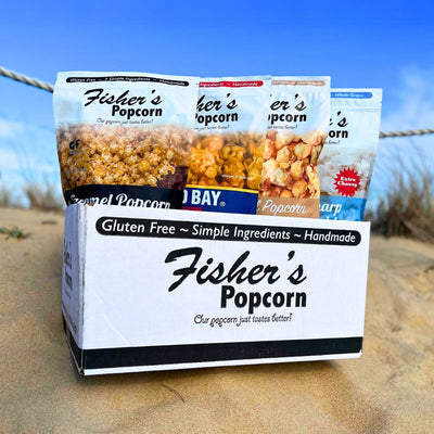 A BYOB 12 Pack of Large Bags sits on sand with a blue sky behind it, featuring a custom popcorn assortment in large resealable bags. The box notes the popcorn is gluten-free, handmade, and made with simple ingredients.