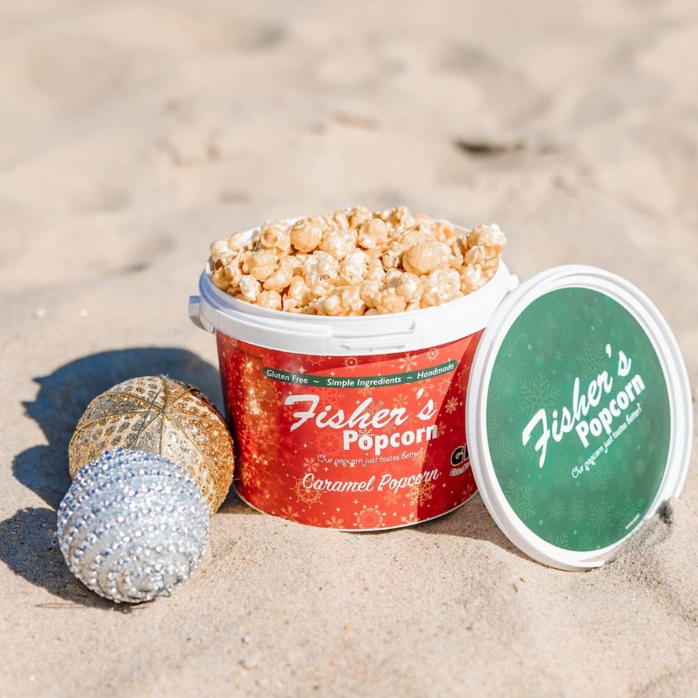 A Small Holiday Tub Fisher’s Popcorn Caramel sits on sandy ground with its lid nearby, joined by gold and silver holiday ornaments beside the gourmet popcorn from Fisher’s Popcorn.