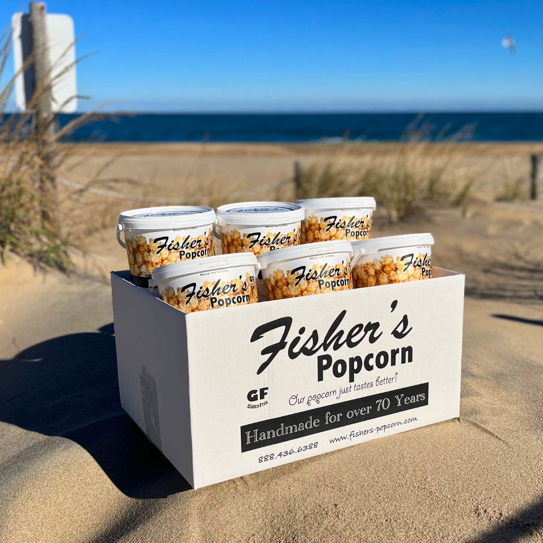 A white box labeled Fisher's Popcorn holds Tubs By The Case (Caramel) and sits on sandy dunes near the beach, with the ocean and blue sky in the background.