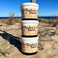Three XXL tubs of BYOB’s 3 Pack are stacked on sandy dunes under a blue sky by the ocean—a perfect popcorn gift for any beach day.