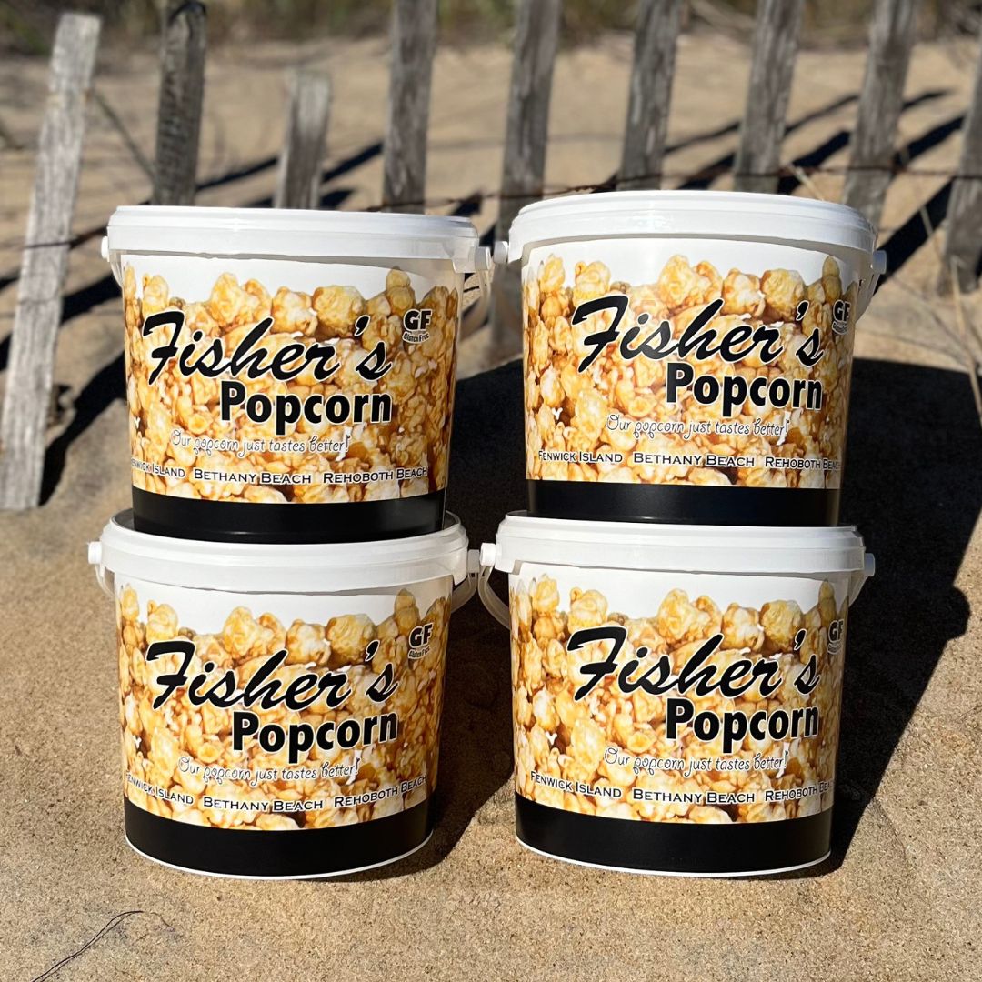 Four BYOB 4 Pack Large Tubs are stacked on sand near a wooden fence and dry grass. Each tub displays caramel popcorn and beach scenes, making this BYOB popcorn gift set the ideal variety pack for any occasion.