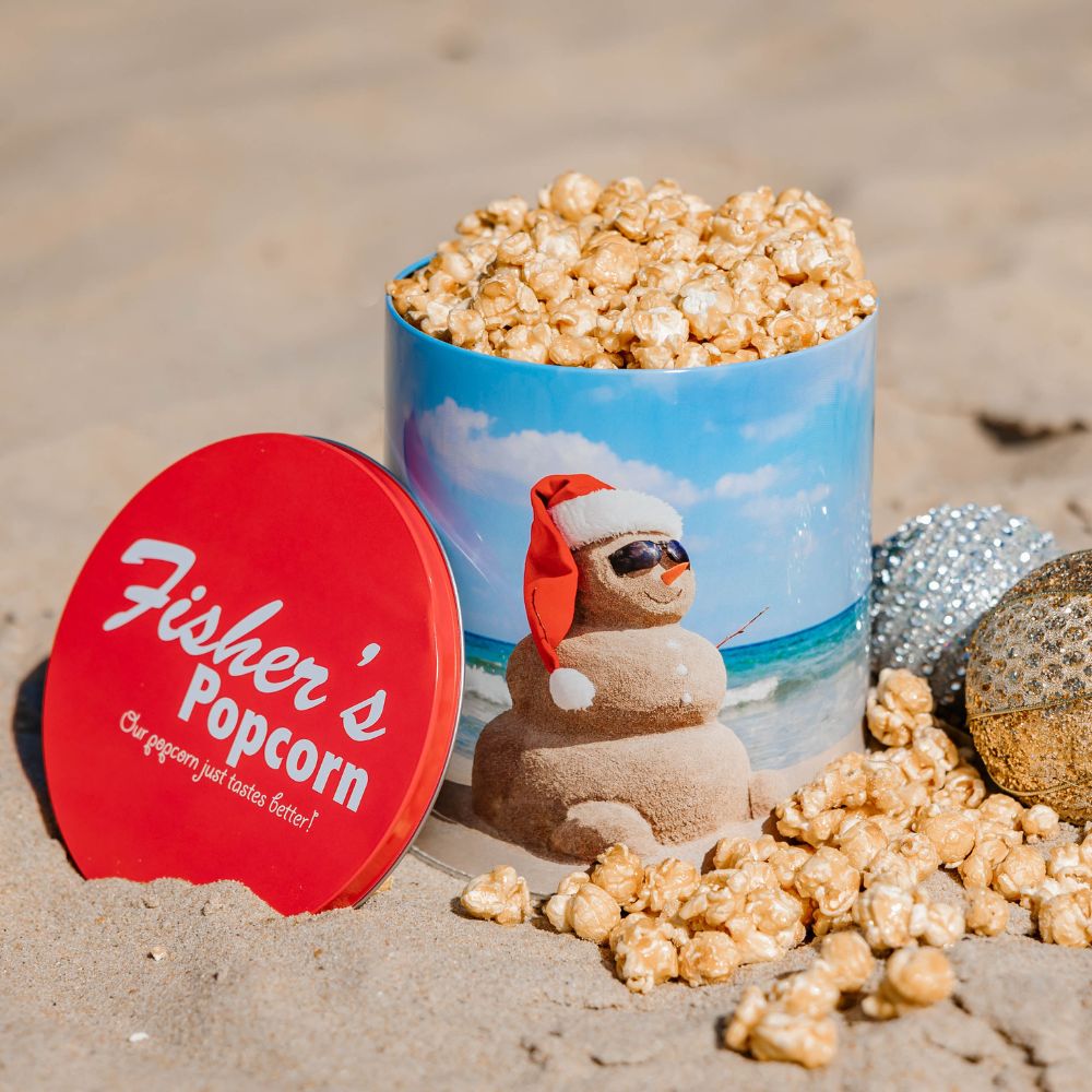 A festive BYOB 6 Pack of 1 Gallon Holiday Tins sits on the sand, with a sunglasses-wearing sand snowman and red lid nearby. Popcorn spills out, making this a perfect addition to any holiday gift bundle.
