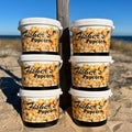 Six BYOB 6 Pack of XL Tubs are stacked on beach sand, with the ocean and clear blue sky behind. Each tub shows bold BYOB branding and caramel popcorn graphics—ideal for your pick 6 popcorn selection!.