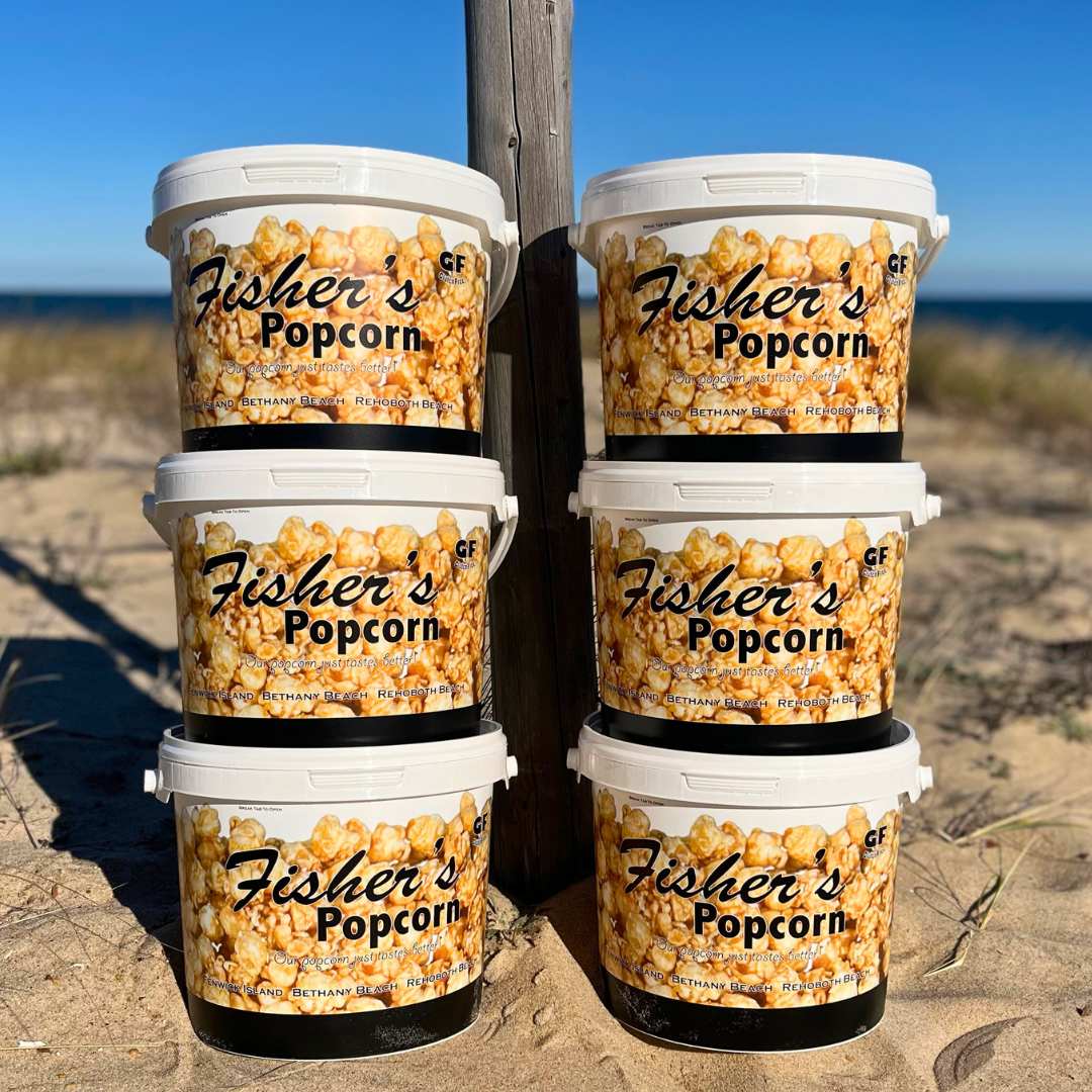 Six BYOB 6 Pack of XL Tubs are stacked on beach sand, with the ocean and clear blue sky behind. Each tub shows bold BYOB branding and caramel popcorn graphics—ideal for your pick 6 popcorn selection!.