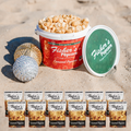 A tub of BYOB Office Party Pack caramel popcorn sits on sand with its lid propped up, two ornaments beside it, and eight smaller caramel popcorn bags in front—perfect as an office gift or festive holiday bundle.