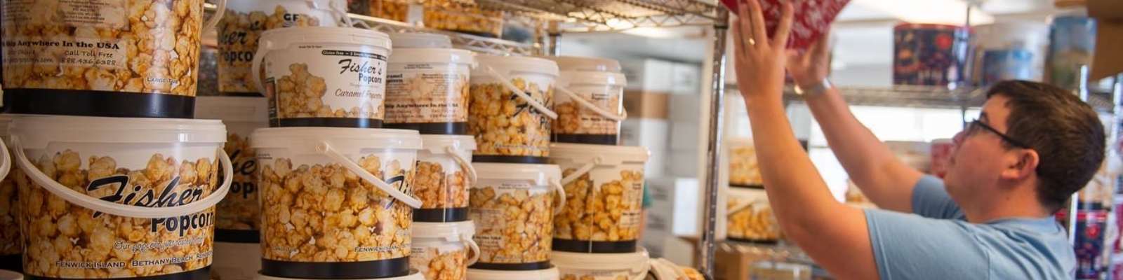 A person wearing glasses and a blue shirt arranges large plastic tubs of popcorn on metal shelves filled with various containers inside a store or storage area.