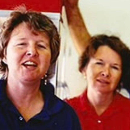 Two women with short brown hair, one in a dark blue top and the other in a red top, stand indoors. The woman in blue is in the foreground, while the other is slightly behind and to the right. A blurred arm is visible in the background.