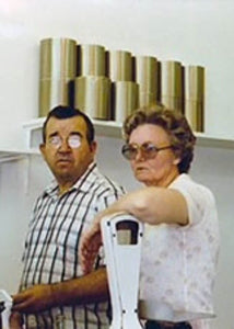A man with a circular white patch over his right eye stands next to a woman in glasses. Both appear serious. Behind them are several cylindrical golden objects arranged on a shelf against a white wall.