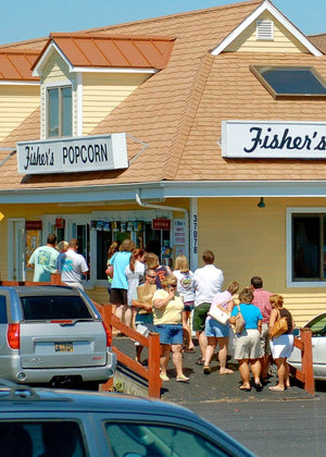 A group of people stand in line outside Fishers Popcorn, a yellow building with white trim. Several cars are parked nearby on a sunny day.