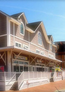 A two-story building with gray siding, white trim, and large windows, featuring a porch with a white railing and stairs. Several storefronts and colorful signs are visible at the ground level.