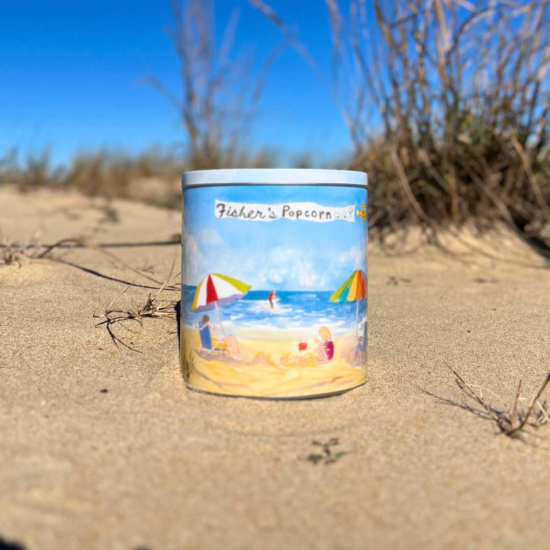 A Fisher’s Popcorn 1 Gallon Beach Tin rests on sandy ground near Fenwick Island, featuring a beach scene and suggesting the fresh popcorn inside.