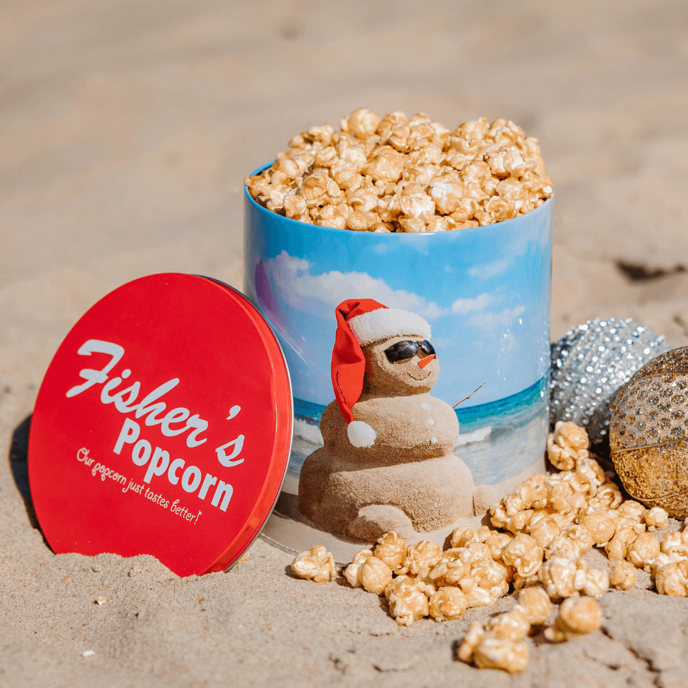 The 1 Gallon Holiday Tin from Fisher's Popcorn, filled with gourmet caramel popcorn, sits on sandy beach. The festive tin shows a sand snowman in sunglasses and a Santa hat, surrounded by popcorn and holiday ornaments.