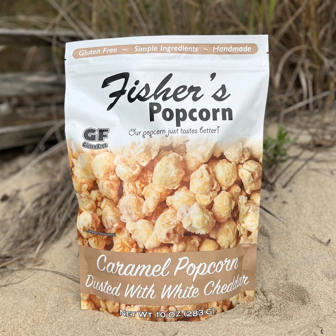 A 10oz bag of Fisher's Popcorn Cheddar Dusted Caramel Popcorn rests on sandy ground with dry grass in the background. The packaging notes that it's gluten free, handmade, and made with simple ingredients.