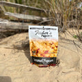A Small Popcorn Bag from Fisher's Popcorn stands upright on sandy dunes with grass and driftwood, evoking a beach vibe. The partially sunlit packaging showcases popcorn, ideal for portion control snacks or unique wedding favors.