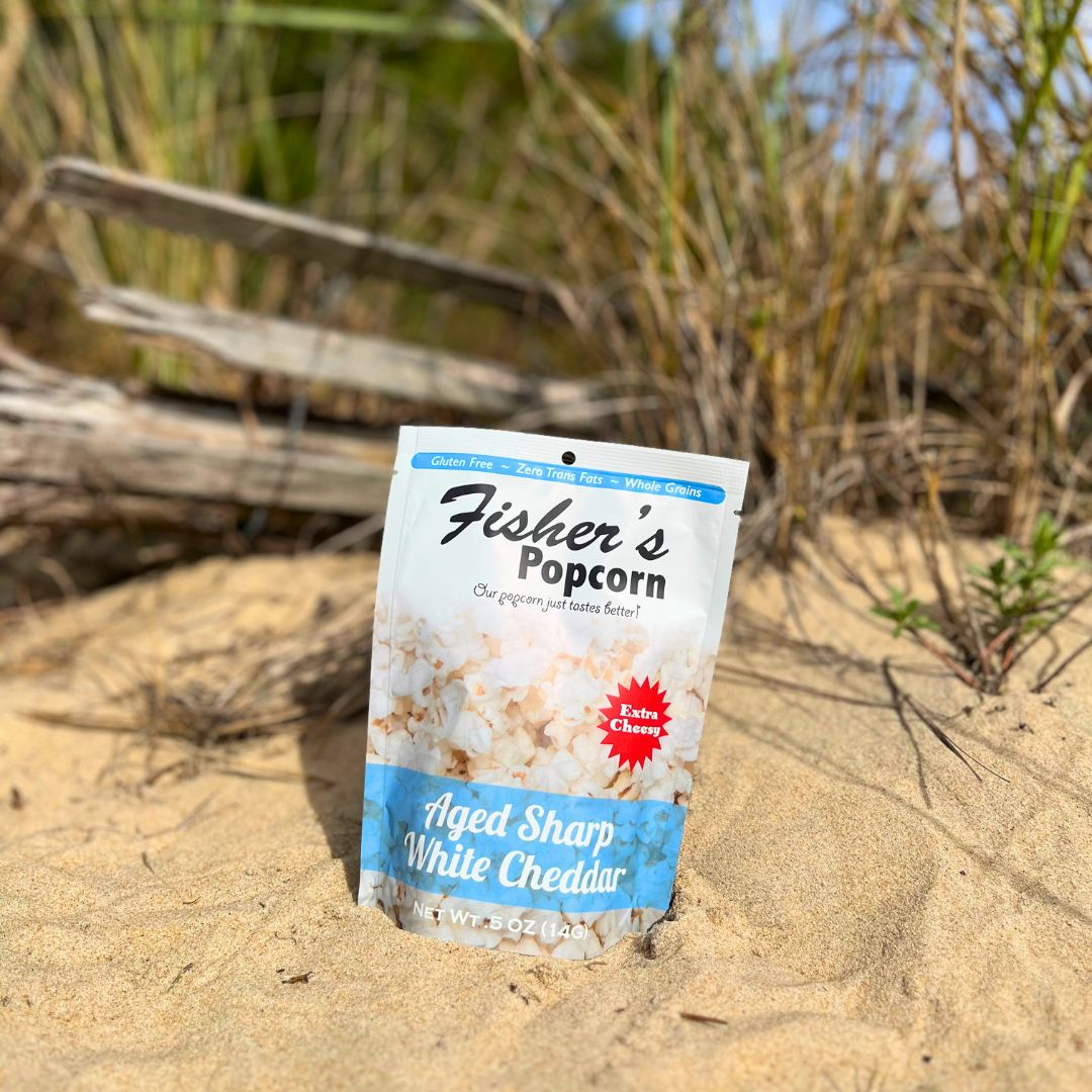 A bag of Fisher's Popcorn Small Popcorn Bags sits upright on sandy ground with grass and wood in the background under bright sunlight—a delicious, portion-controlled snack ideal for wedding favors.