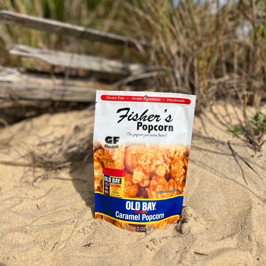 A Small Popcorn Bag from Fisher's Popcorn stands upright in sandy soil, making a unique wedding favor, with grass and a weathered wooden fence softly blurred in the background.