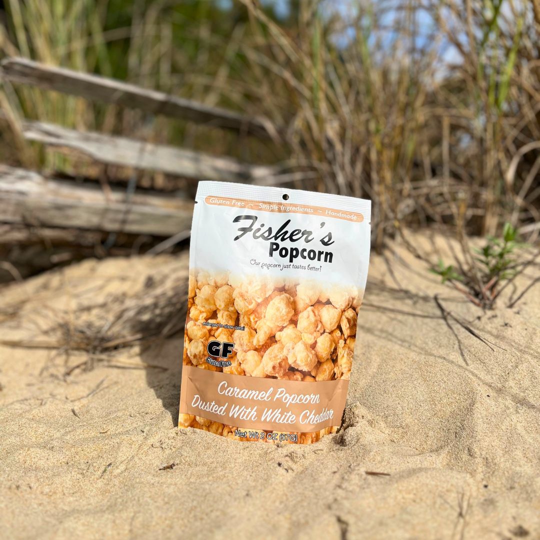 A Fisher’s Popcorn Small Popcorn Bag, caramel flavor dusted with white cheddar, stands upright on sandy ground—perfect as tasty wedding favors or portion-control snacks for your next event.