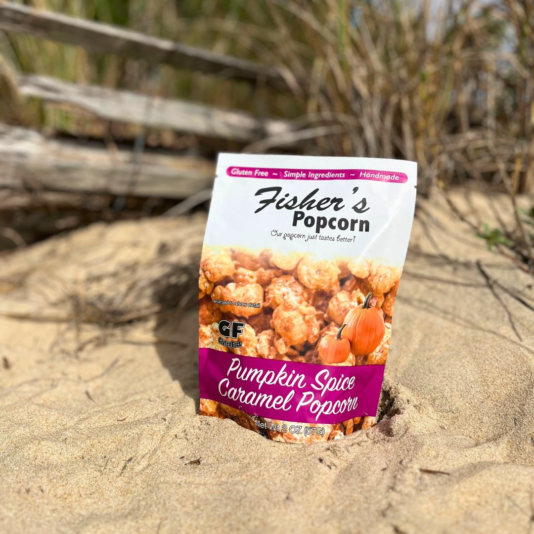 A bag of Fisher's Popcorn Small Popcorn Bags rests on the beach, making it an ideal choice for wedding favors or portion-controlled snacks, with dried grass and a wooden fence in the background.