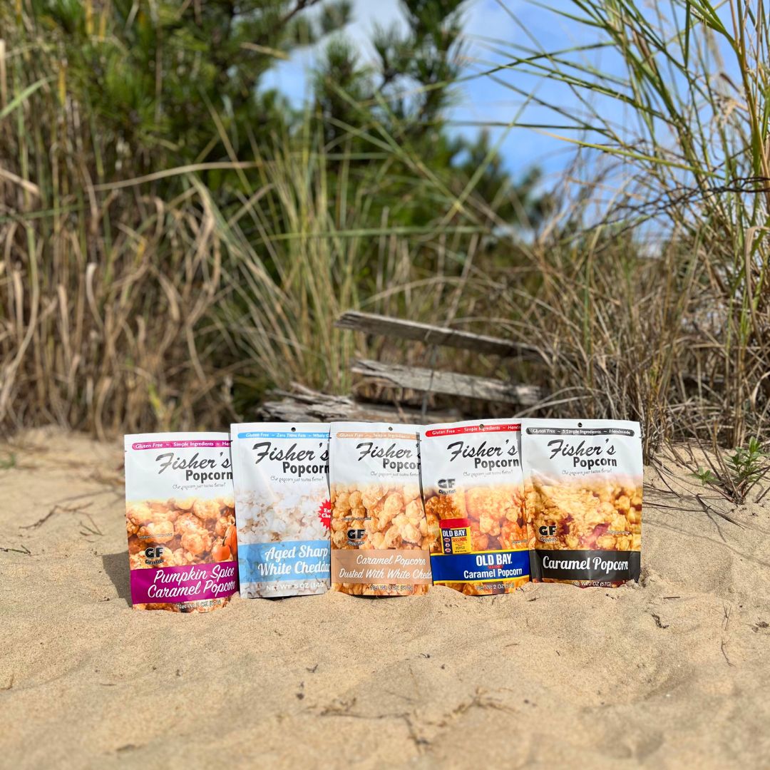 Four Small Popcorn Bags from Fisher's Popcorn are arranged on sandy ground with tall grass in the background, creating a coastal vibe—ideal for portion control snacks or as unique wedding favors.