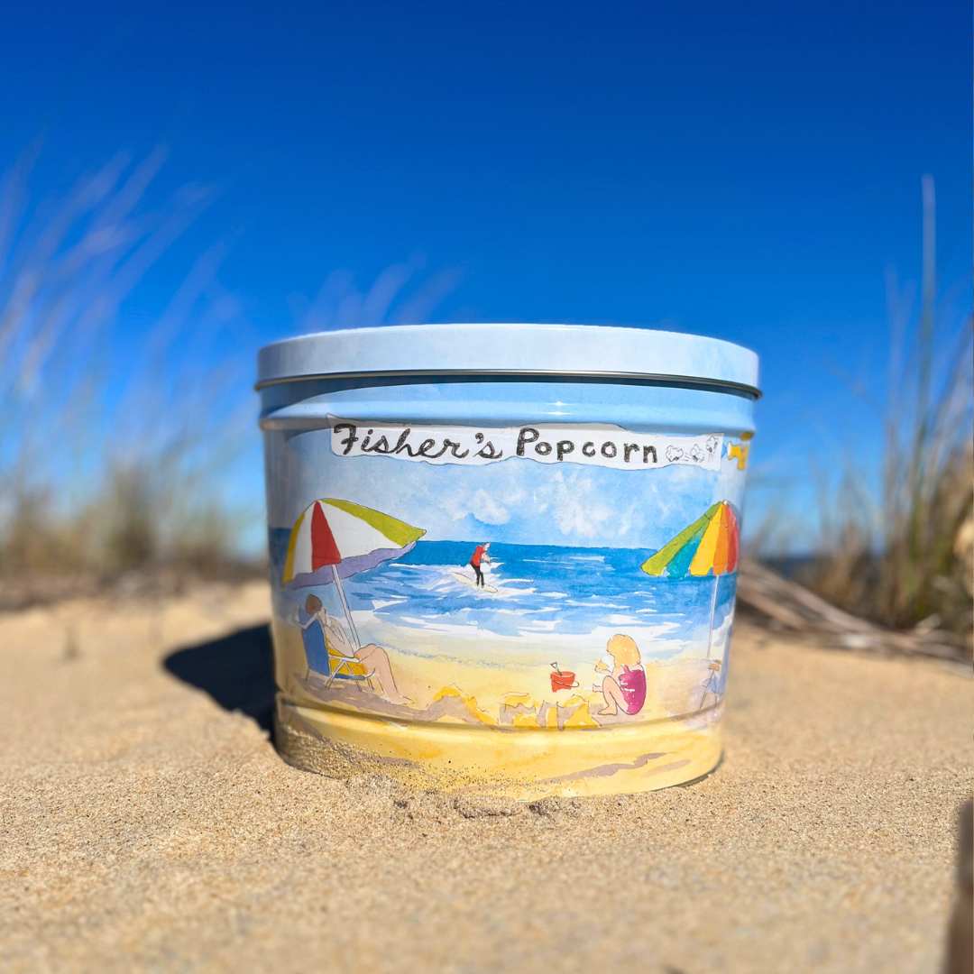 A Fisher's Popcorn 2 Gallon Beach Tin sits on sandy ground with dry grass and a clear blue sky in the background, making it perfect for family gifts or sharing delicious popcorn by the shore.
