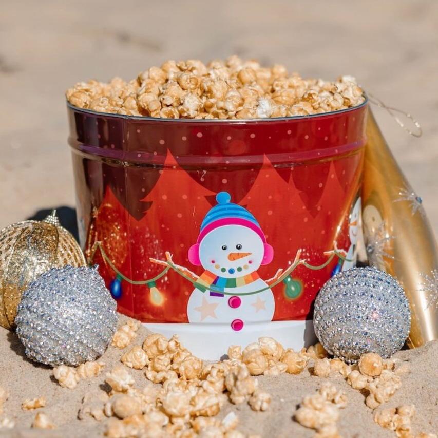 The Fisher's Popcorn 2 Gallon Holiday Tin features a festive snowman design and comes filled with caramel popcorn—an ideal seasonal gift, pictured on sandy ground with gold and silver ornaments and scattered popcorn.