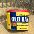 A 2 Gallon OLD BAY® Tin of Fisher's Popcorn sits on the sand, partially in sunlight, with grassy dunes blurred in the background.