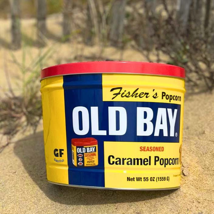 A 2 Gallon OLD BAY® Tin of Fisher's Popcorn sits on the sand, partially in sunlight, with grassy dunes blurred in the background.