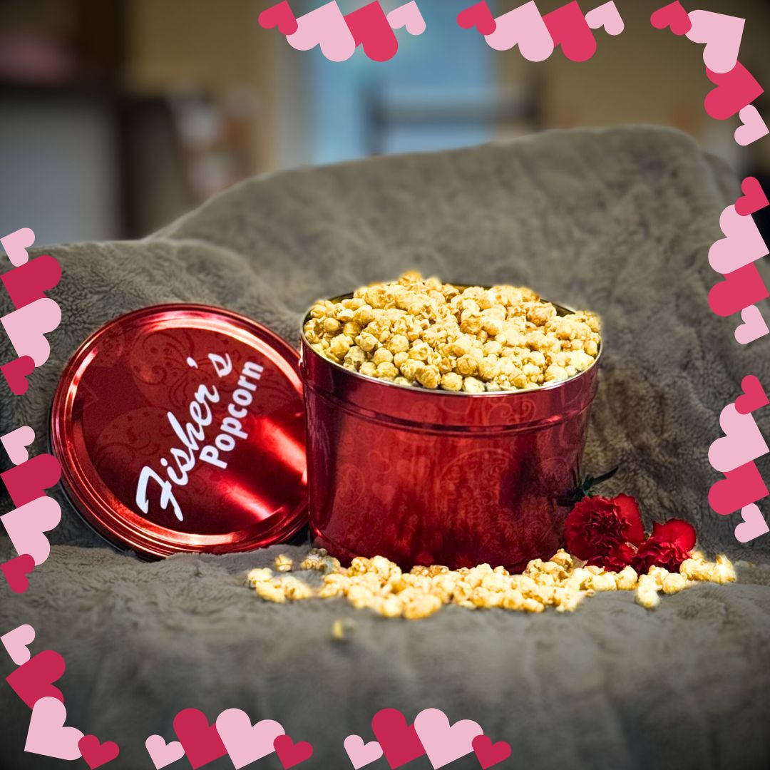 An open 2 Gallon Valentine Tin from Fisher's Popcorn sits on a gray blanket, filled with gourmet caramel popcorn spilling out. Two red carnations are nearby, and the scene is bordered with pink and red heart shapes.
