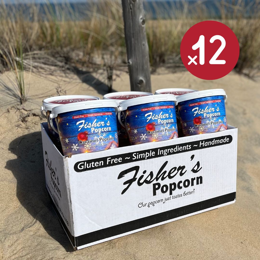 A box of Fisher's Popcorn 2023 Christmas Tub 12 Packs sits on sandy ground near beach grass, with a red ×12 icon—an ideal holiday gift that's gluten free, handmade, and crafted with simple ingredients.