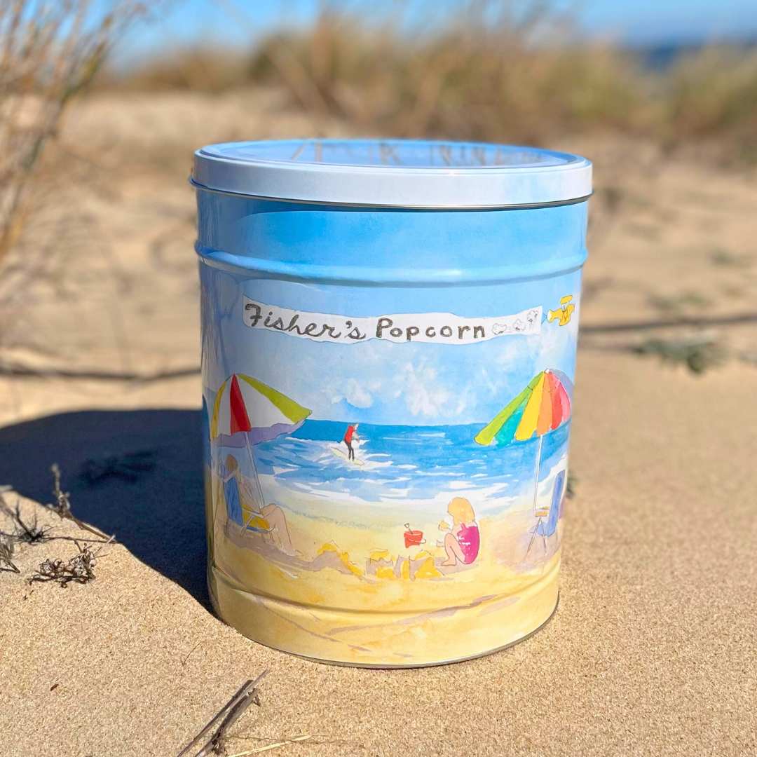 Fisher’s Popcorn 3.5 Gallon Beach Tin features a colorful beach scene with umbrellas, a child, and the ocean on sandy dunes under a blue sky—making it the perfect popcorn gift.