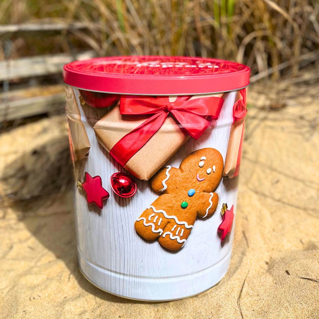 The Fisher's Popcorn 3.5 Gallon Holiday Tin, featuring festive images and a red lid, rests on the sand with grassy plants and a wooden fence in the background—making it the perfect holiday gift.