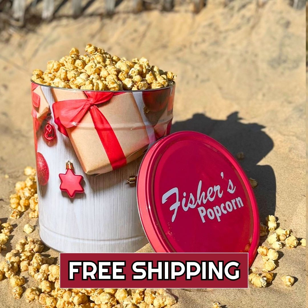 The 3.5 Gallon Holiday Tin by Fisher's Popcorn, featuring a festive gift design, sits on sand with gourmet popcorn spilling out and its red lid beside it. A banner announces FREE SHIPPING.