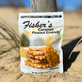 A bag of Fisher's Popcorn Caramel Peanut Crunch stands upright on sandy ground, with grass and a blurred blue sky behind it. The package displays stacked pieces of caramel peanut brittle candy.