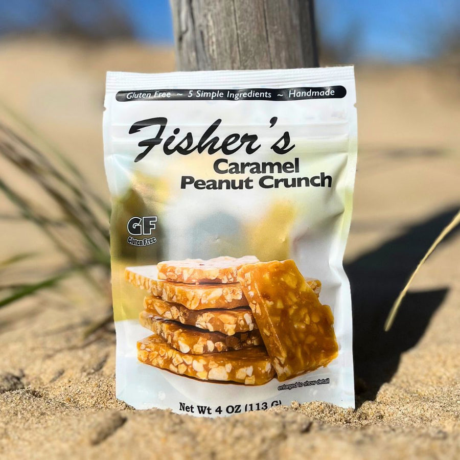 A bag of Fisher's Popcorn Caramel Peanut Crunch stands upright on sandy ground, with grass and a blurred blue sky behind it. The package displays stacked pieces of caramel peanut brittle candy.