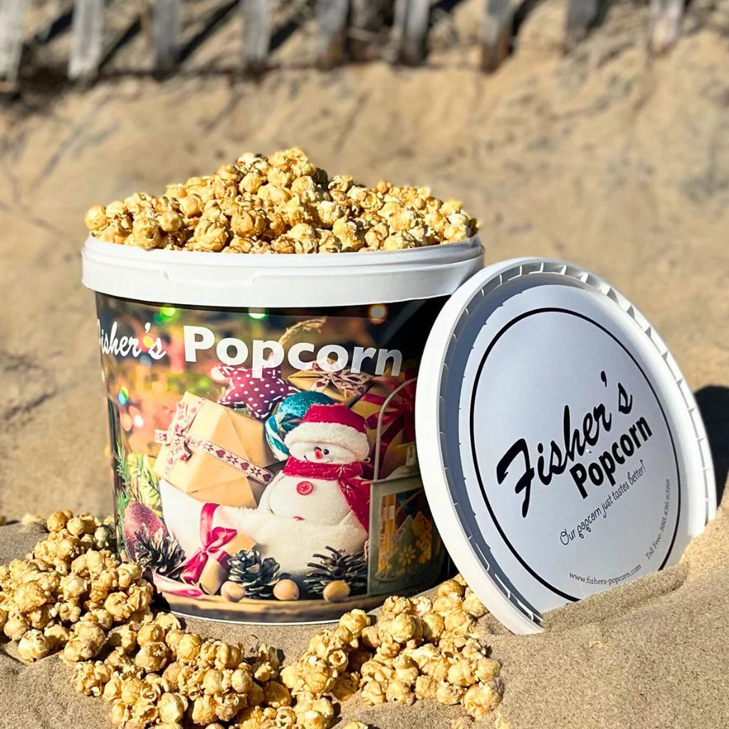 A large Fisher's Popcorn Christmas in July Tub sits on the sand with caramel popcorn spilling out. Decorated with gift boxes and a snowman, its lid nearby displays the logo—perfect for gifting and comes with free shipping!.